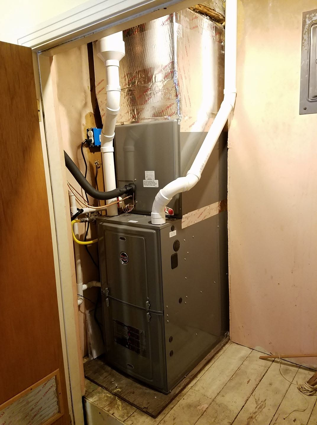 Furnace and HVAC unit in a narrow utility closet with exposed ductwork and pipes.
