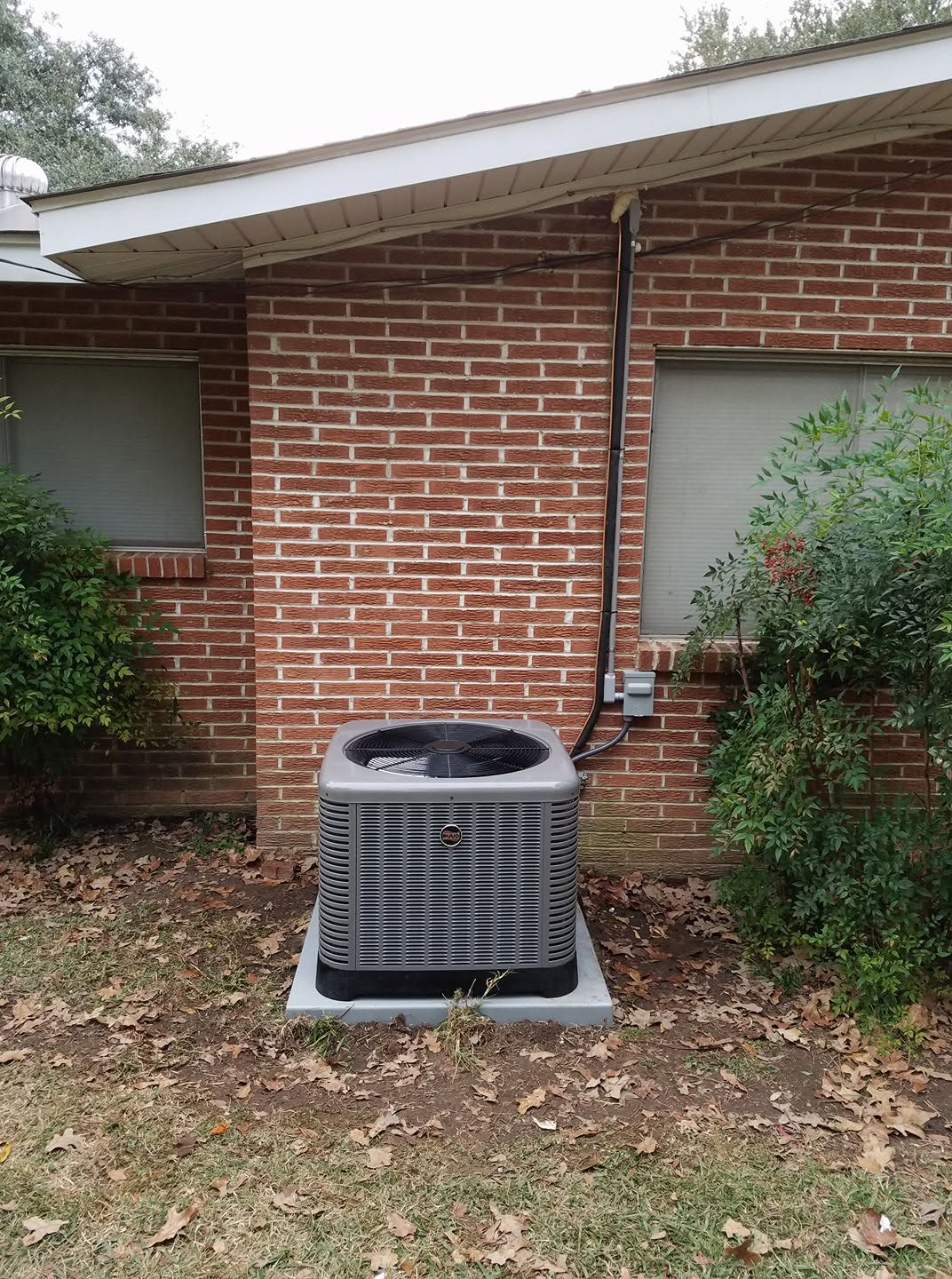 Central air conditioner unit beside a red brick house with bushes and fallen leaves.
