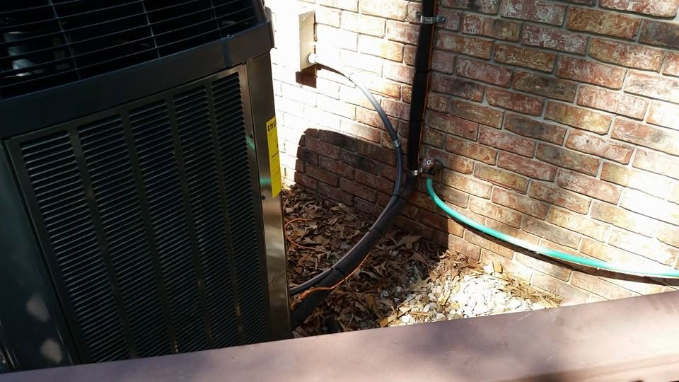 Outdoor HVAC unit beside a brick wall, with hoses and fallen leaves in a shaded corner.