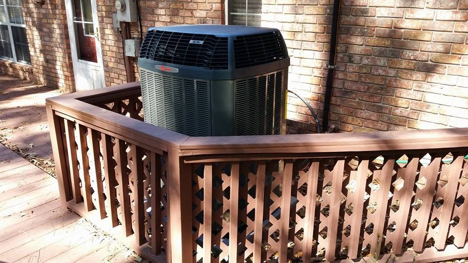 Outdoor air conditioner unit on a wooden deck beside a brick house and railing.