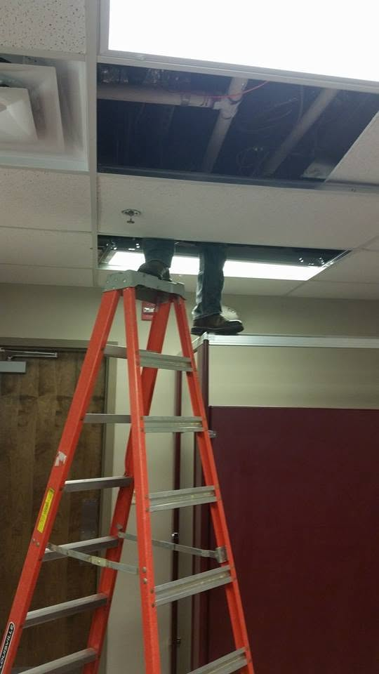 Person on a ladder working in an open ceiling tile above an office room