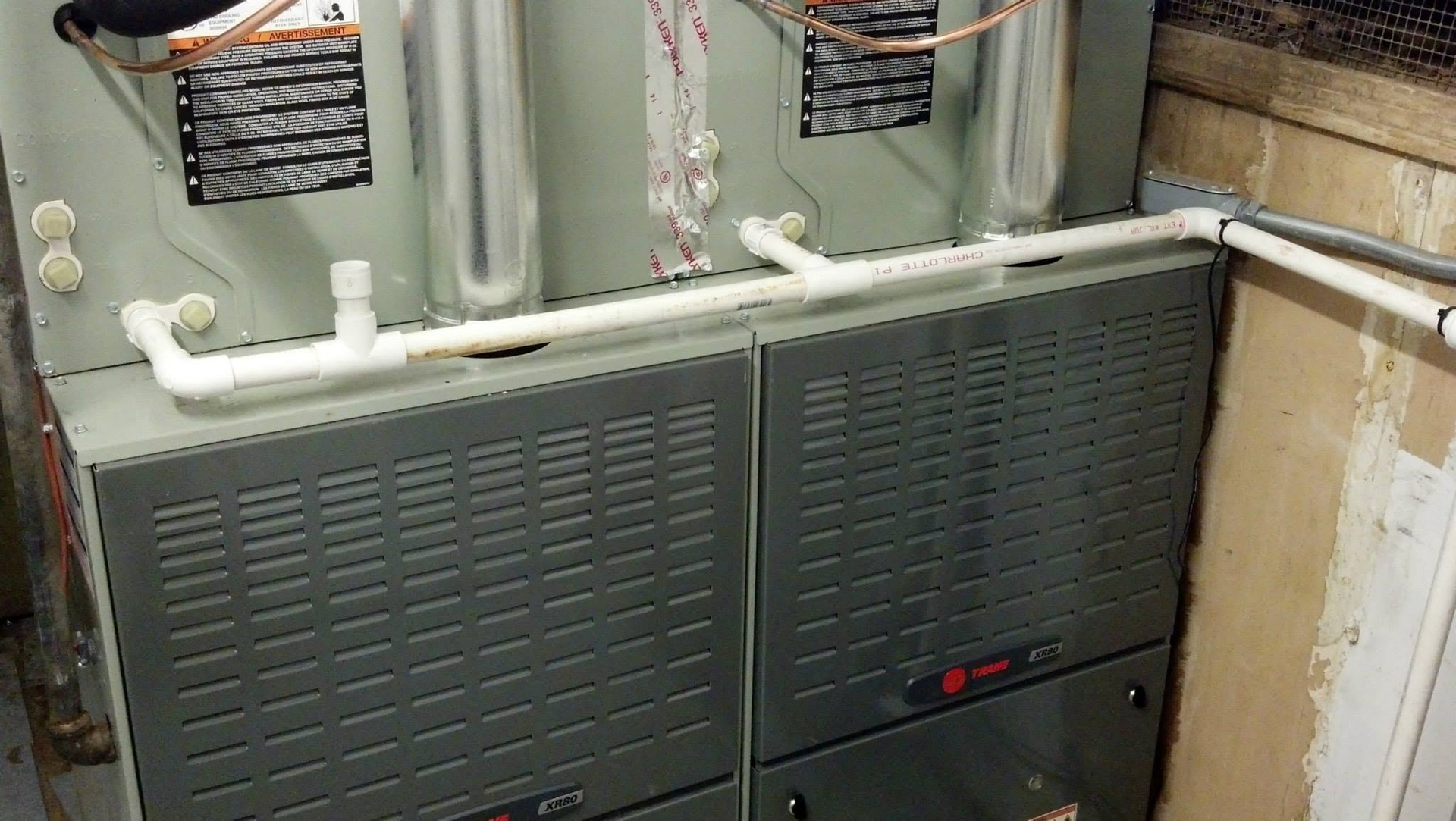 Gray HVAC furnace and ductwork in a utility closet with pipes and vents above.