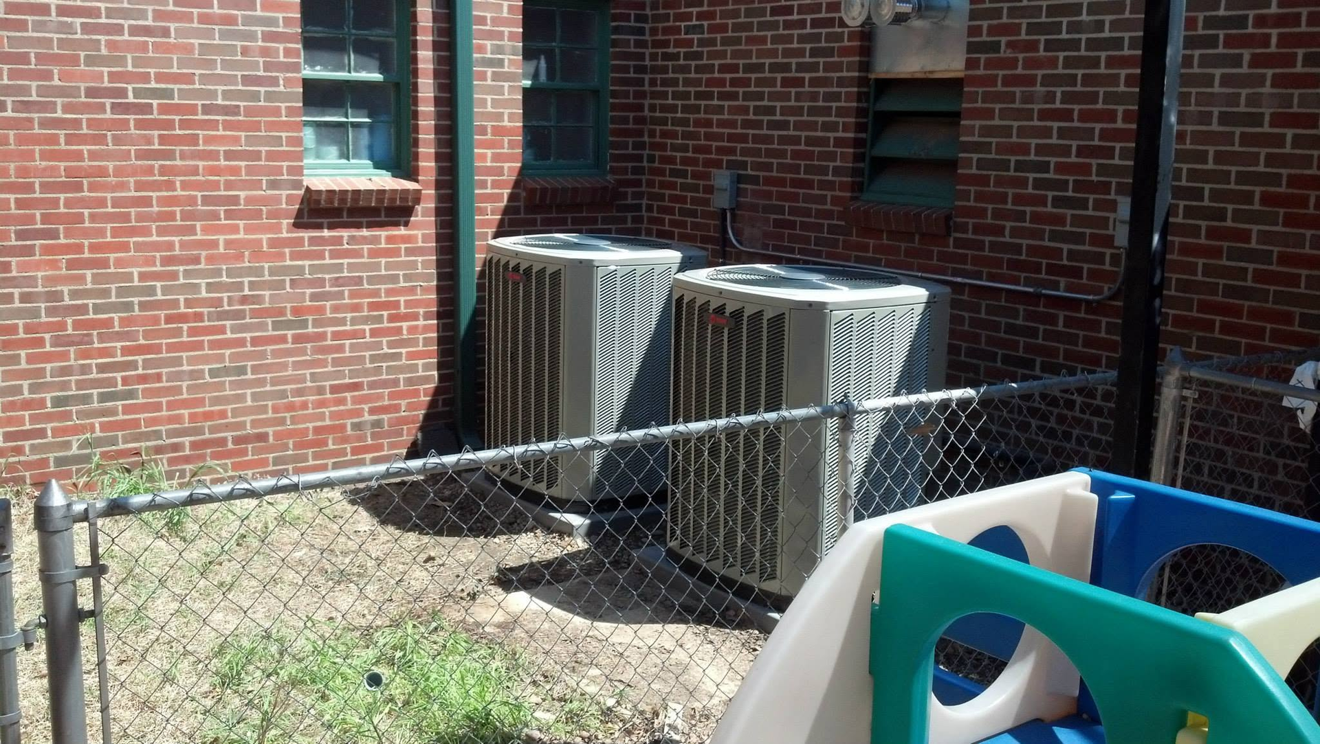 Two outdoor HVAC units beside a brick building, with a chain-link fence and plastic playground equipment in the foreground.