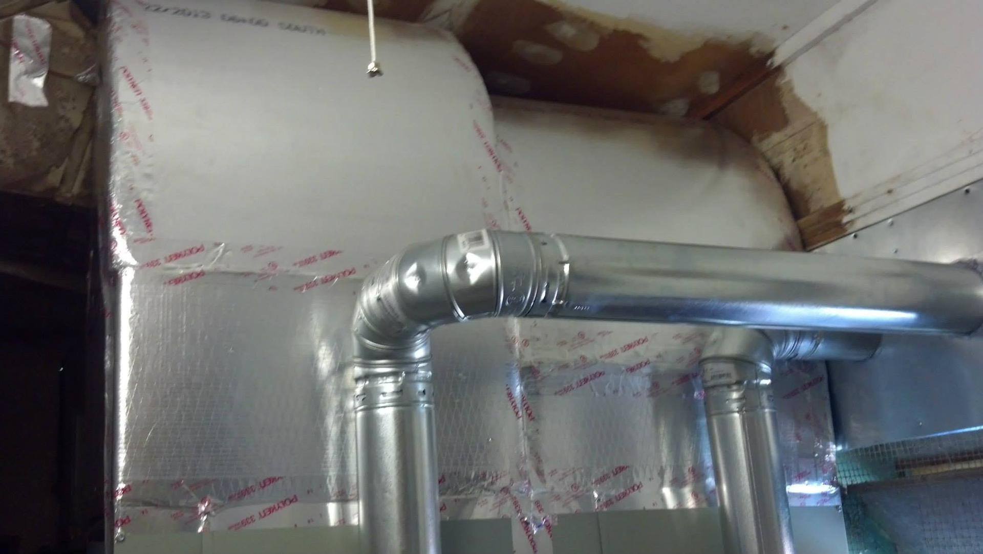 Insulated HVAC ductwork and pipes running through a basement ceiling with exposed framing and foil insulation.