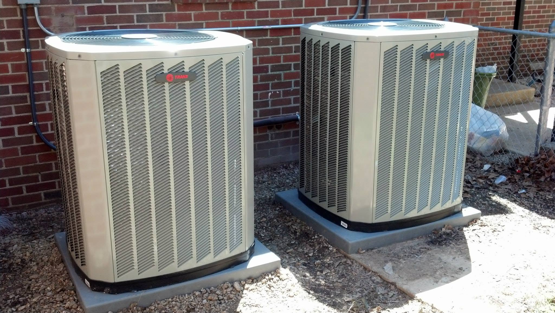 Two outdoor air conditioner units beside a brick wall, one beige and one gray, on a concrete pad.