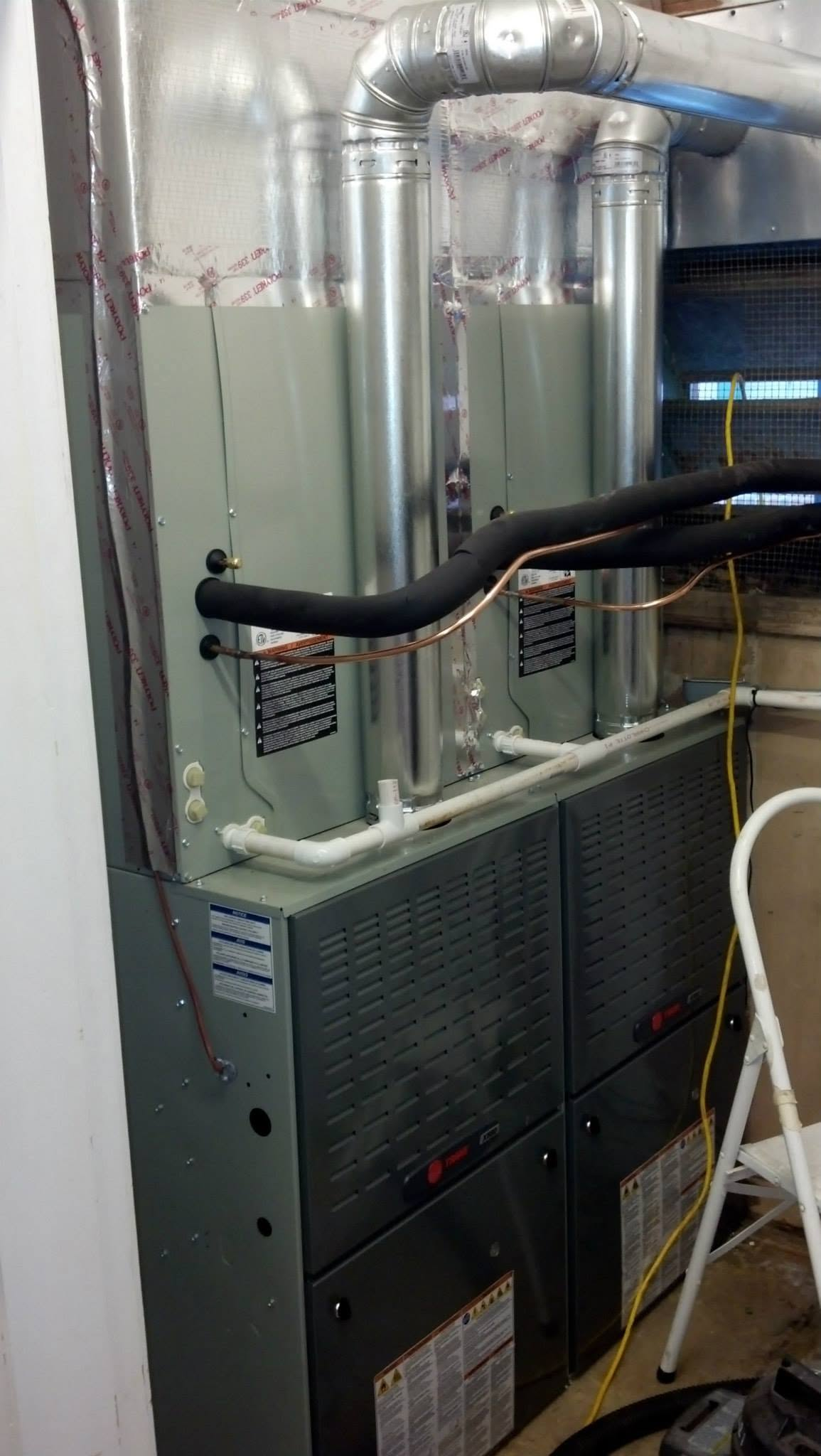 Home furnace with silver ducts, black hoses, and vented gray unit in a utility room.