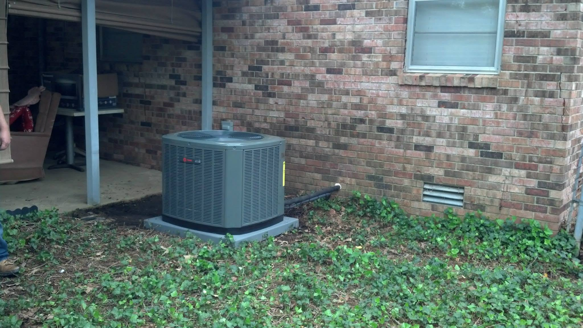 Outdoor HVAC unit beside a brick house wall, with grass and leaves in the yard.