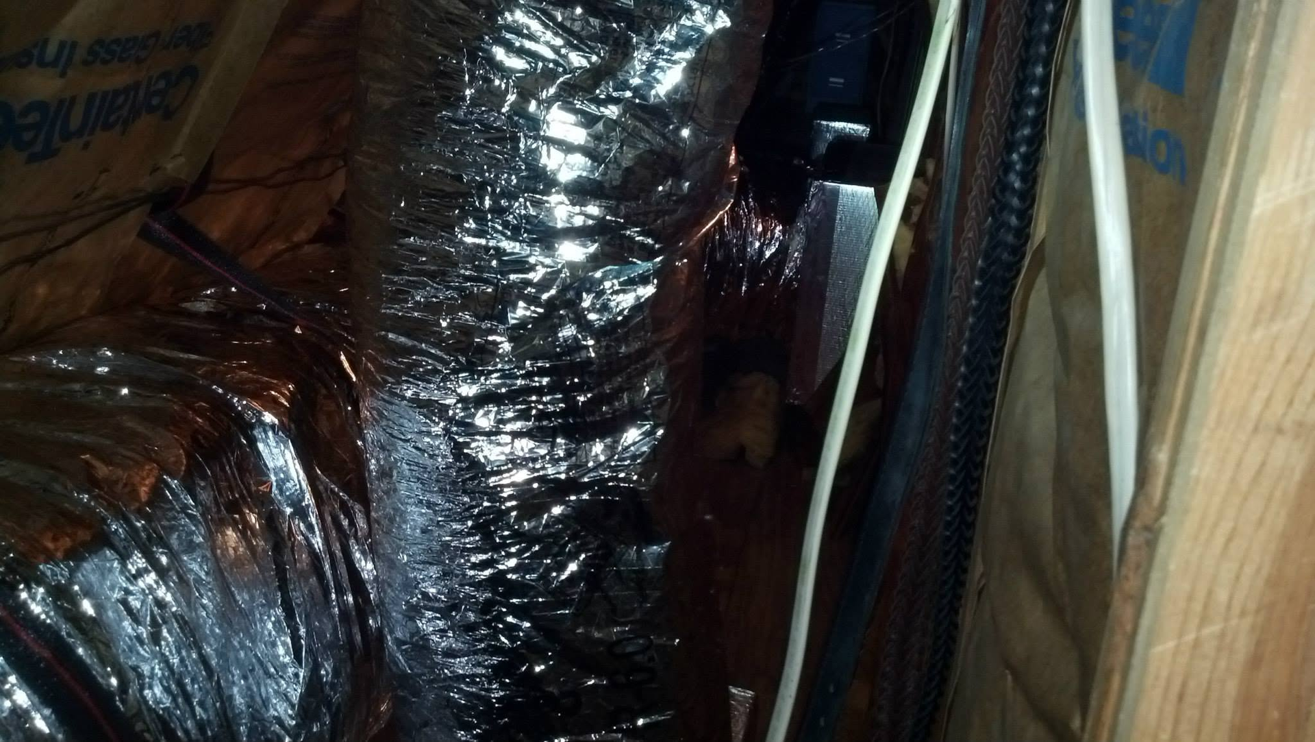 Black insulated HVAC ducts and a wooden wall inside an unfinished attic or crawl space.