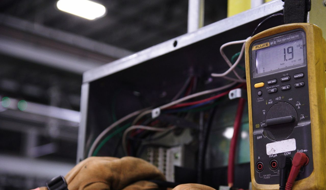Gloved hands use a multimeter on exposed wires inside an electrical panel.