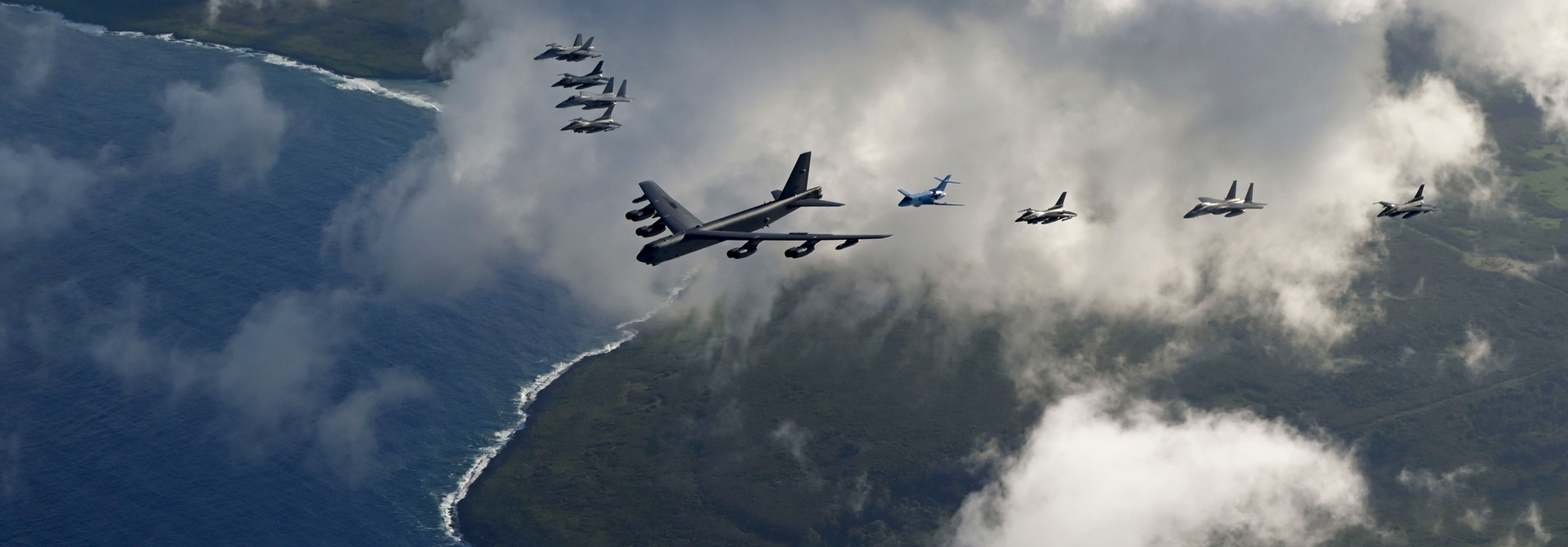 Military aircraft flying in formation over a cloudy blue sky, with a large transport plane leading.