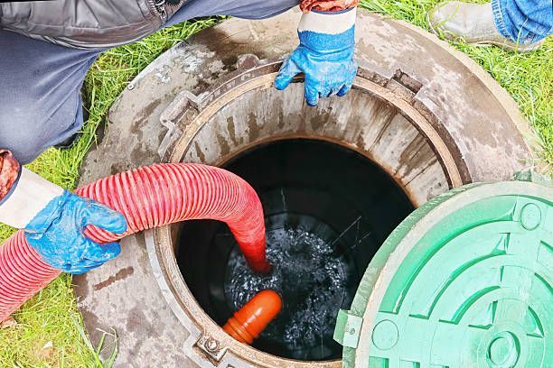 Man in blue gloves pumping liquid from a sewage access point with a red hose and open green lid.