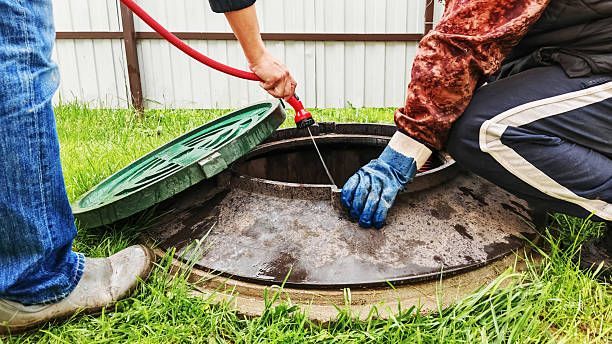 Two people near an open septic tank, one holding a hose, the other with a gloved hand inside.