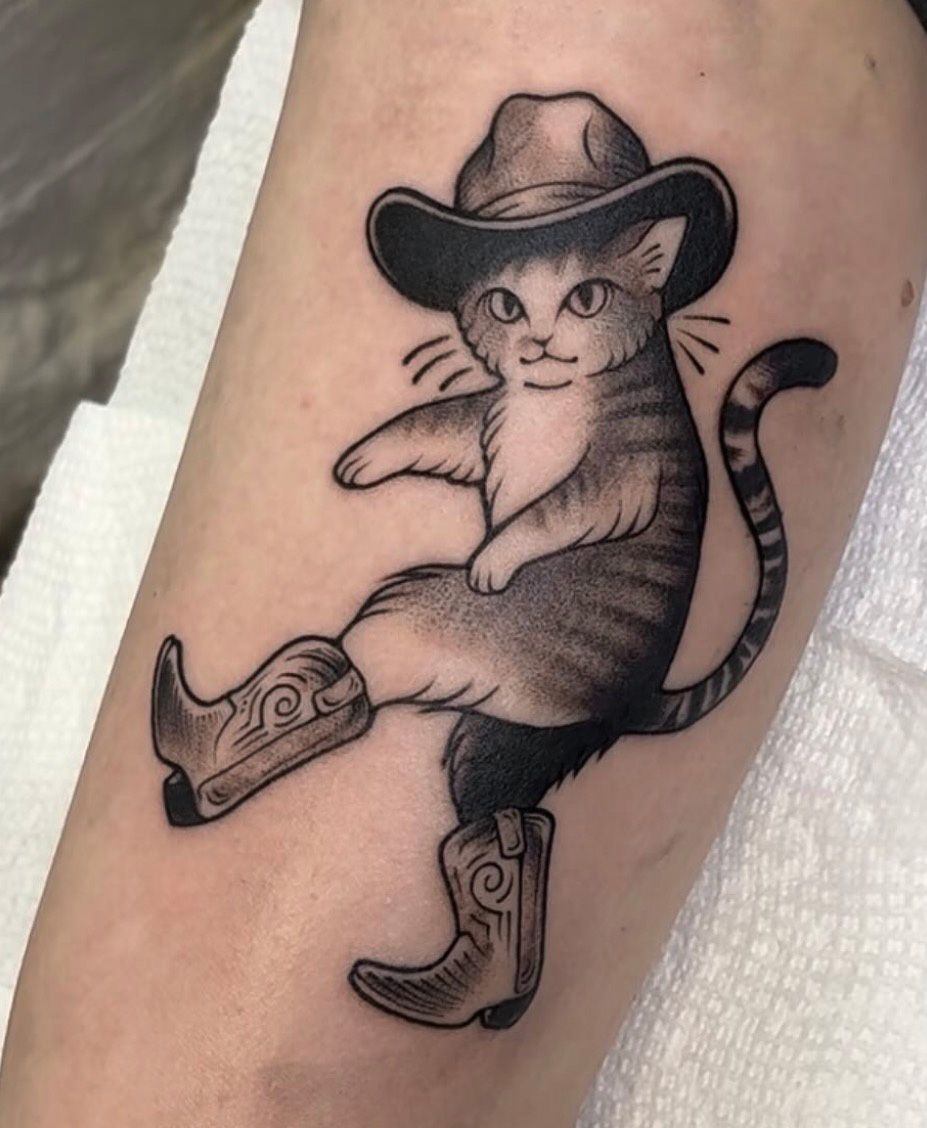 Black and gray illustrative tattoo by Jacy Andrew featuring a playful tabby cat wearing a cowboy hat and western boots, with detailed stippled shading.