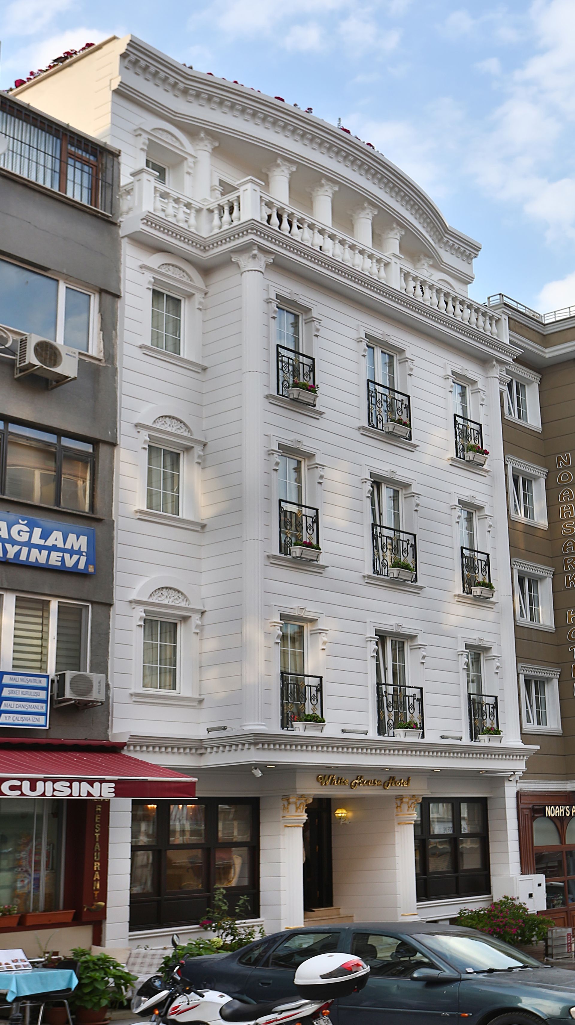 White ornate hotel building with balconies and shops at street level, parked cars in front