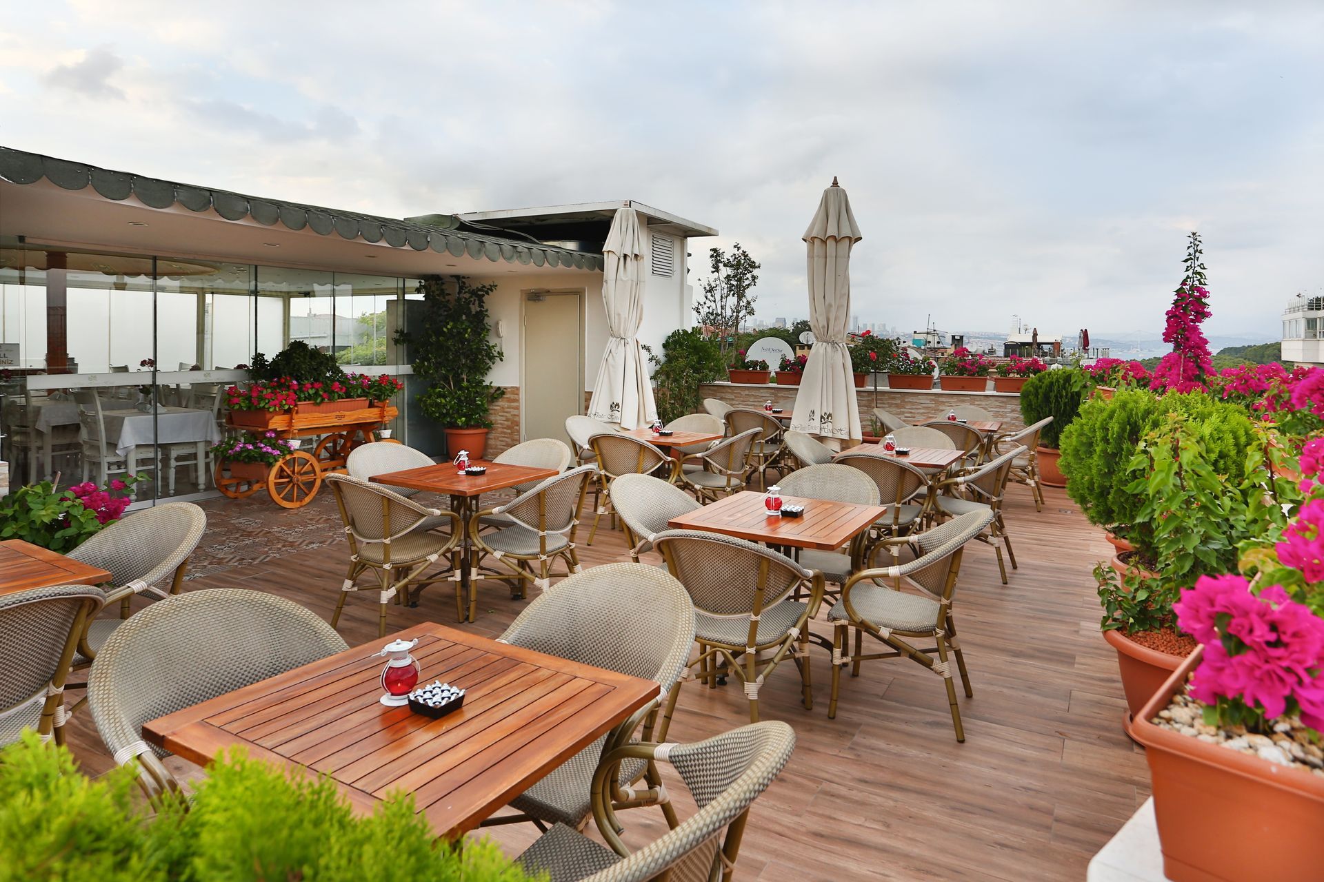Outdoor rooftop café with wicker chairs, wooden tables, and bright pink flowers overlooking a city view