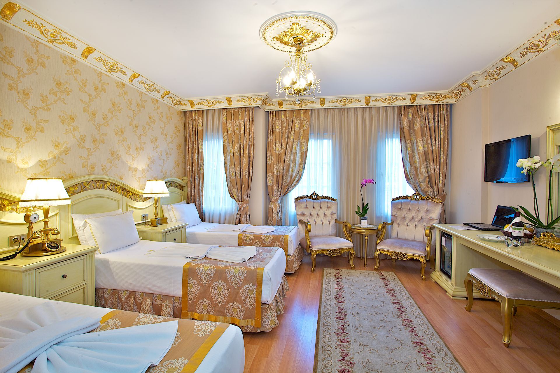 Elegant hotel room with two beds, ornate gold decor, chandelier, curtains, and TV