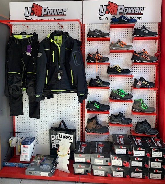 Esposizione di abbigliamento da lavoro e scarpe antinfortunistiche U-Power in un negozio. Abbigliamento, scarpe e scatole su scaffali rossi e pannelli forati.