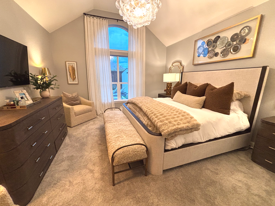 Cozy beige bedroom with a bed, dresser, armchair, large window, and chandelier