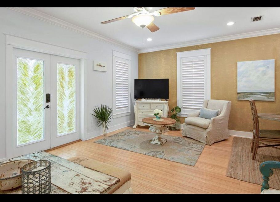 Bright living room with white French doors, beige chairs, TV, rug, and soft coastal decor.