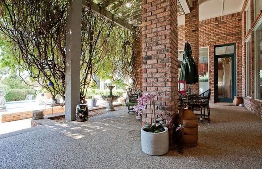 Covered brick porch with potted flowers, wicker chairs, and hanging moss beside a tree-lined yard