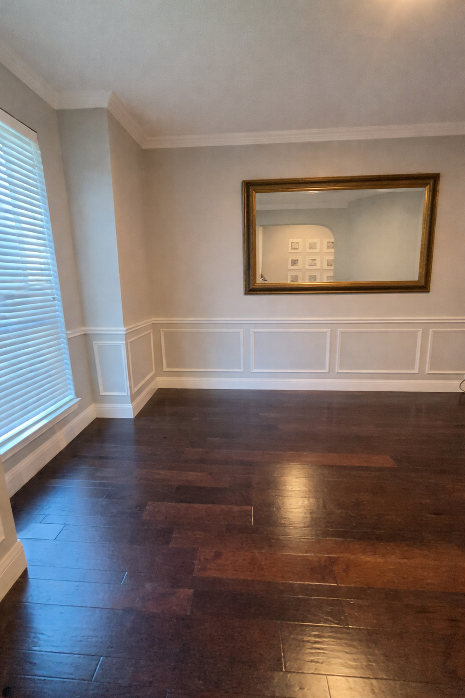 Empty dining room with dark hardwood floors, white wainscoting, and a large framed mirror on the wall.