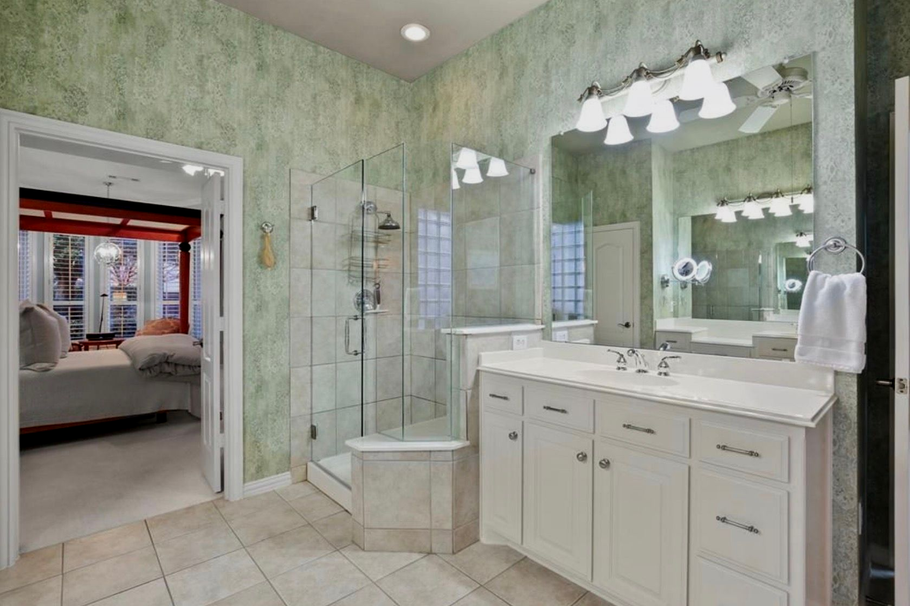 Spacious beige bathroom with white vanity, large mirror, glass shower, and open doorway to a bedroom.