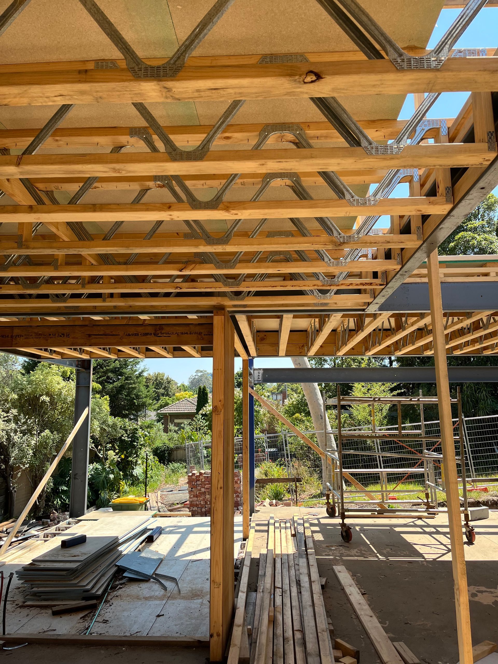 Construction site: wooden beams and steel supports form a partially constructed roof structure. Sunlight streams through.