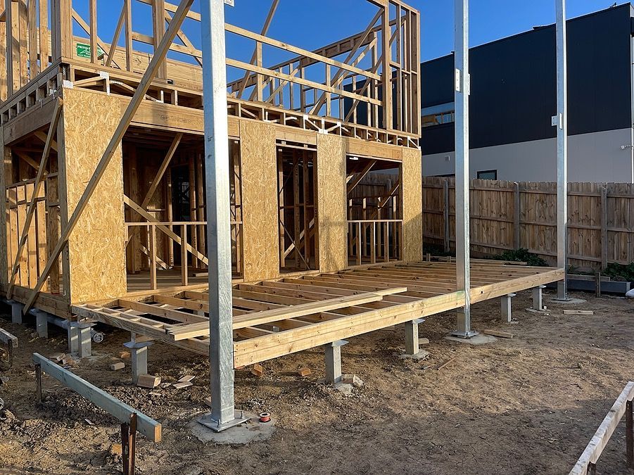 Wooden house under construction; exposed framing, deck, and support columns; outdoor setting.