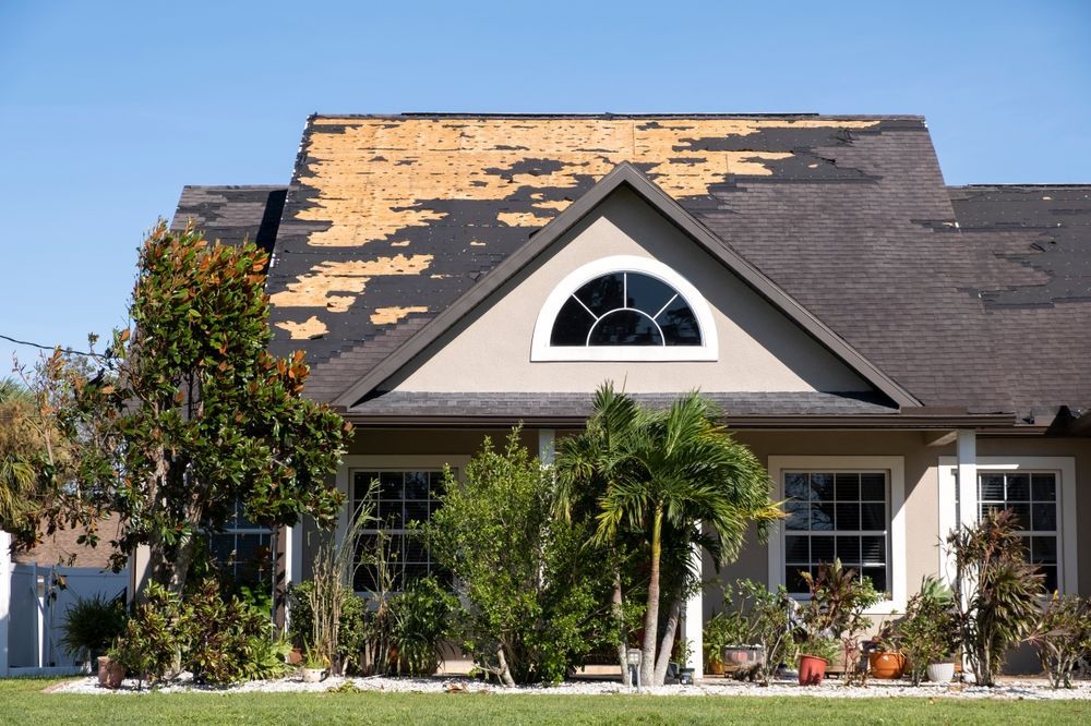 House with damaged roof; shingles missing, exposing underlayment. Sunny day.
