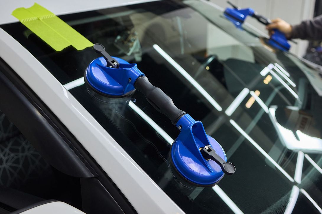 Persona usando ventosas azules para levantar un parabrisas. Coche blanco en un garaje iluminado.