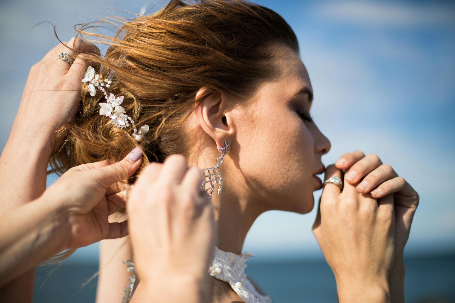 Bride having hair styled, ocean in background. Hands adjust hair and rings.
