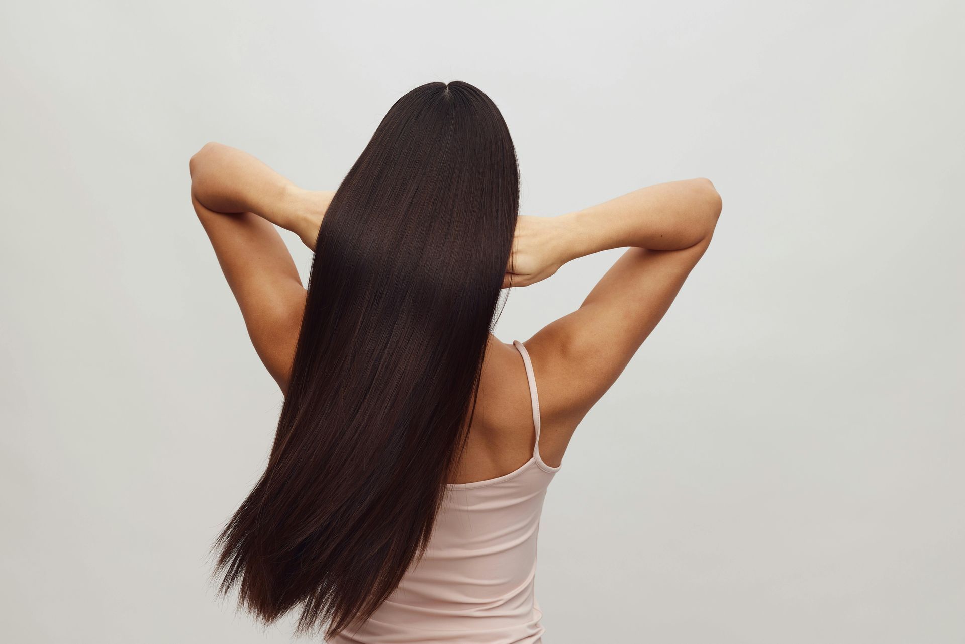 Woman with long, shiny, straight, dark hair, arms raised behind head, wearing a light pink tank top.