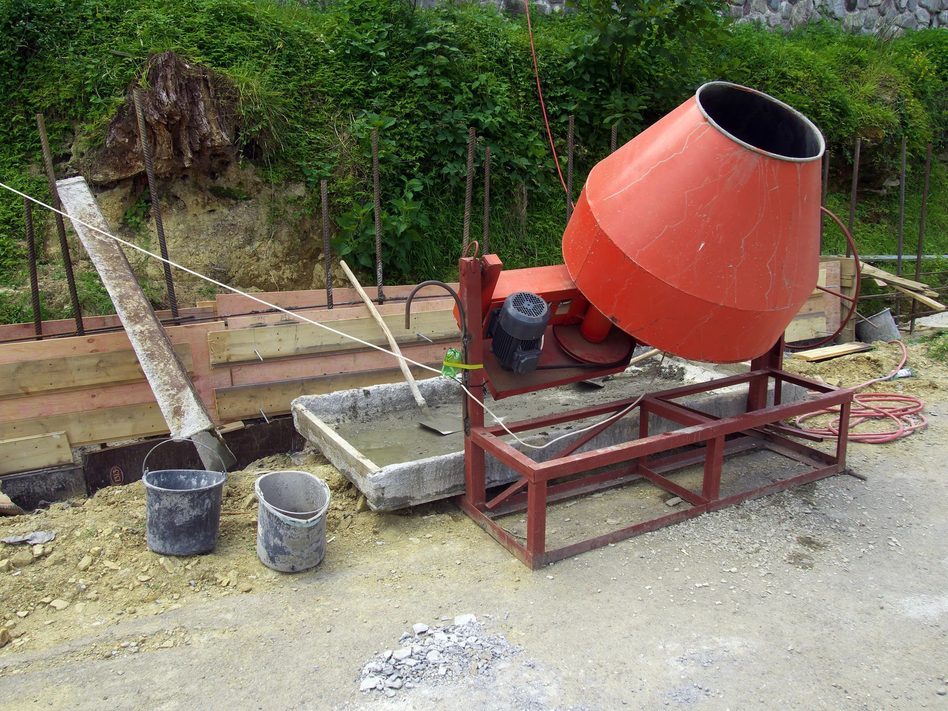 Una hormigonera naranja sobre un soporte metálico junto a cubos y una canaleta en una obra en construcción.