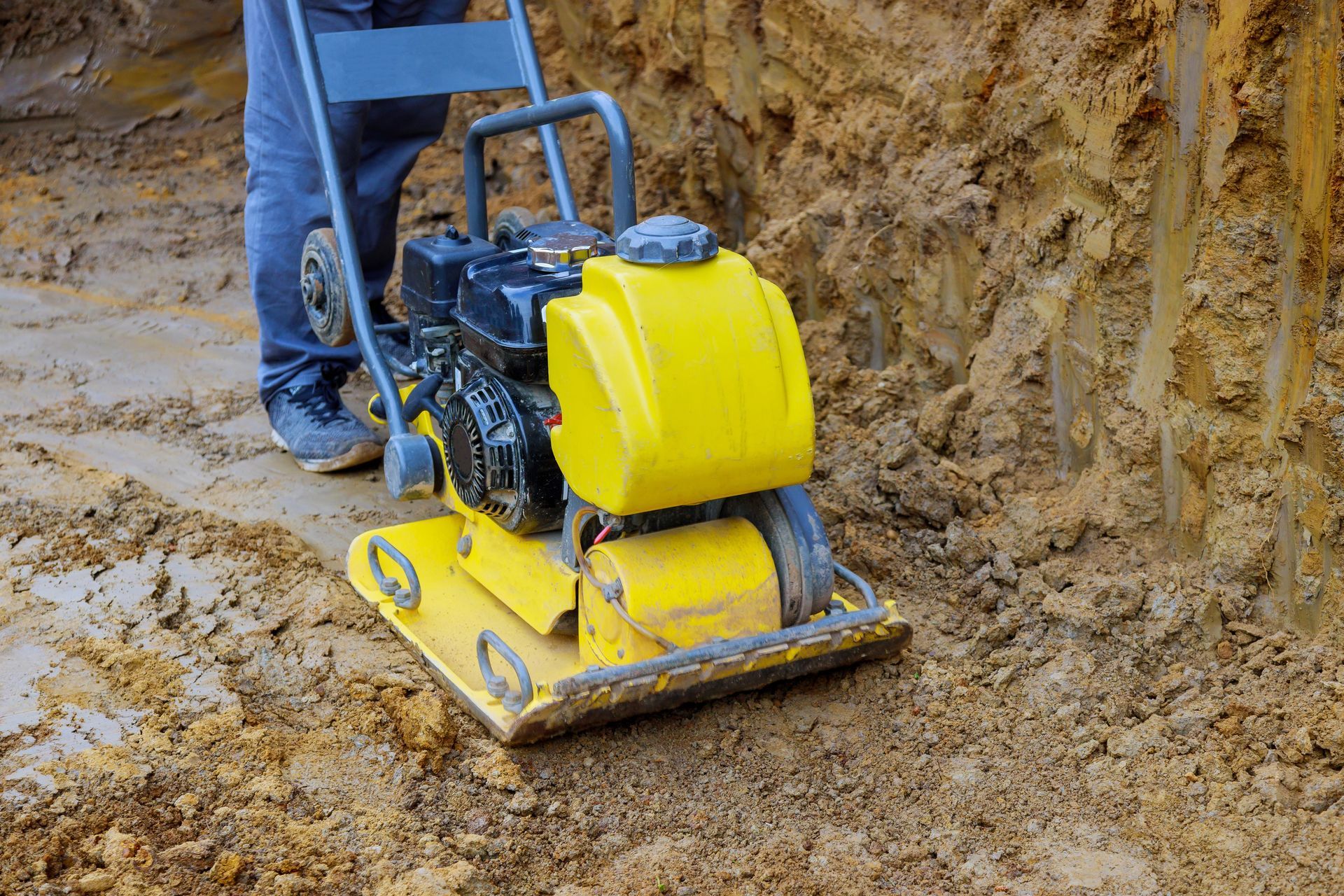 Obrero manejando una compactadora de placas amarillas en terreno fangoso en una obra de construcción.
