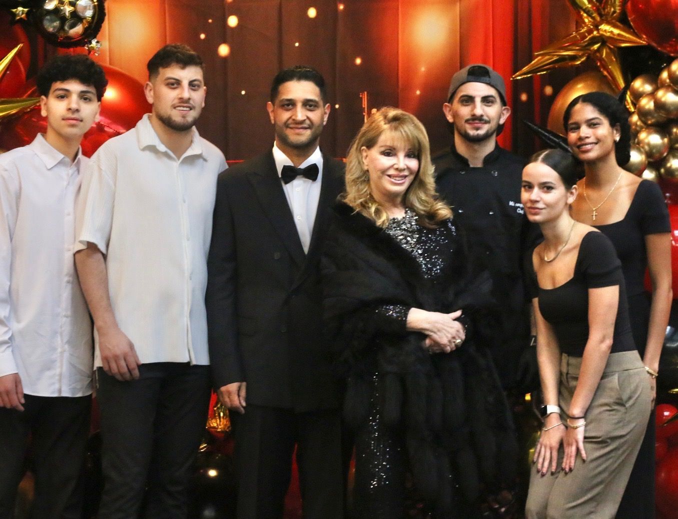 Group of seven people posing formally in front of a decorated backdrop with warm lights.
