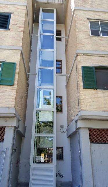 Vano ascensore verticale in vetro sulla facciata di un condominio beige con persiane verdi