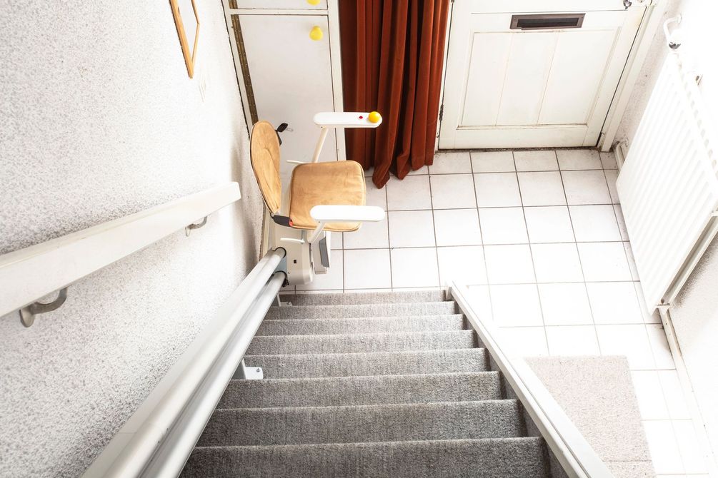 Stretto vano scale interno con moquette grigia, piastrelle bianche, ringhiere e una piccola sedia di legno sul pianerottolo