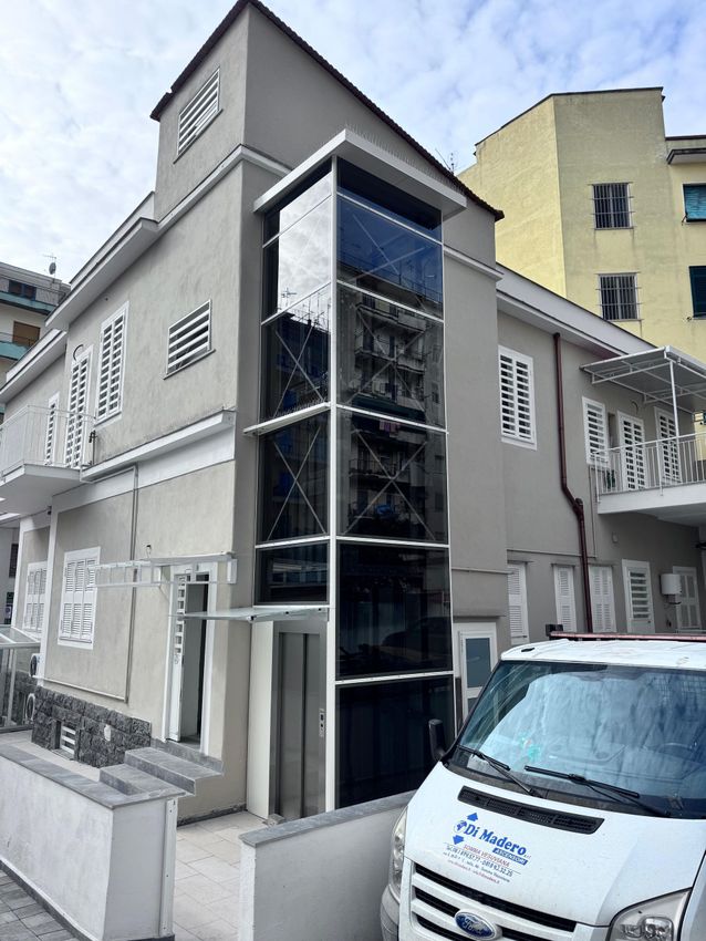 Edificio grigio a più piani con vano ascensore in vetro, rifiniture bianche e un furgone bianco parcheggiato davanti