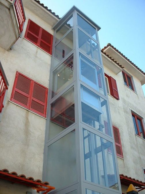 Ascensore in vetro addossato a un edificio bianco con persiane rosse e tetto in tegole