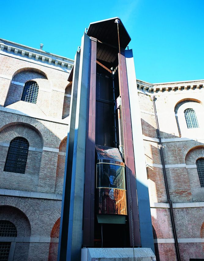 Edificio storico in mattoni con un alto vano centrale in vetro per l’ascensore, contro un cielo azzurro
