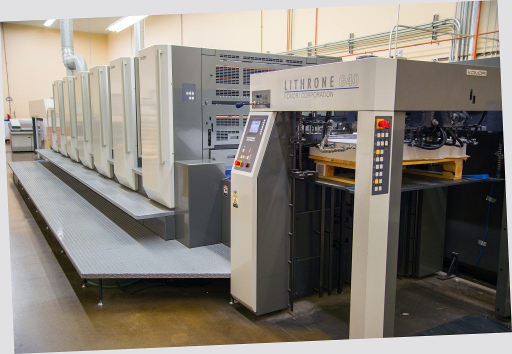 Large industrial printing press in a factory, with gray machinery and a row of printing units.