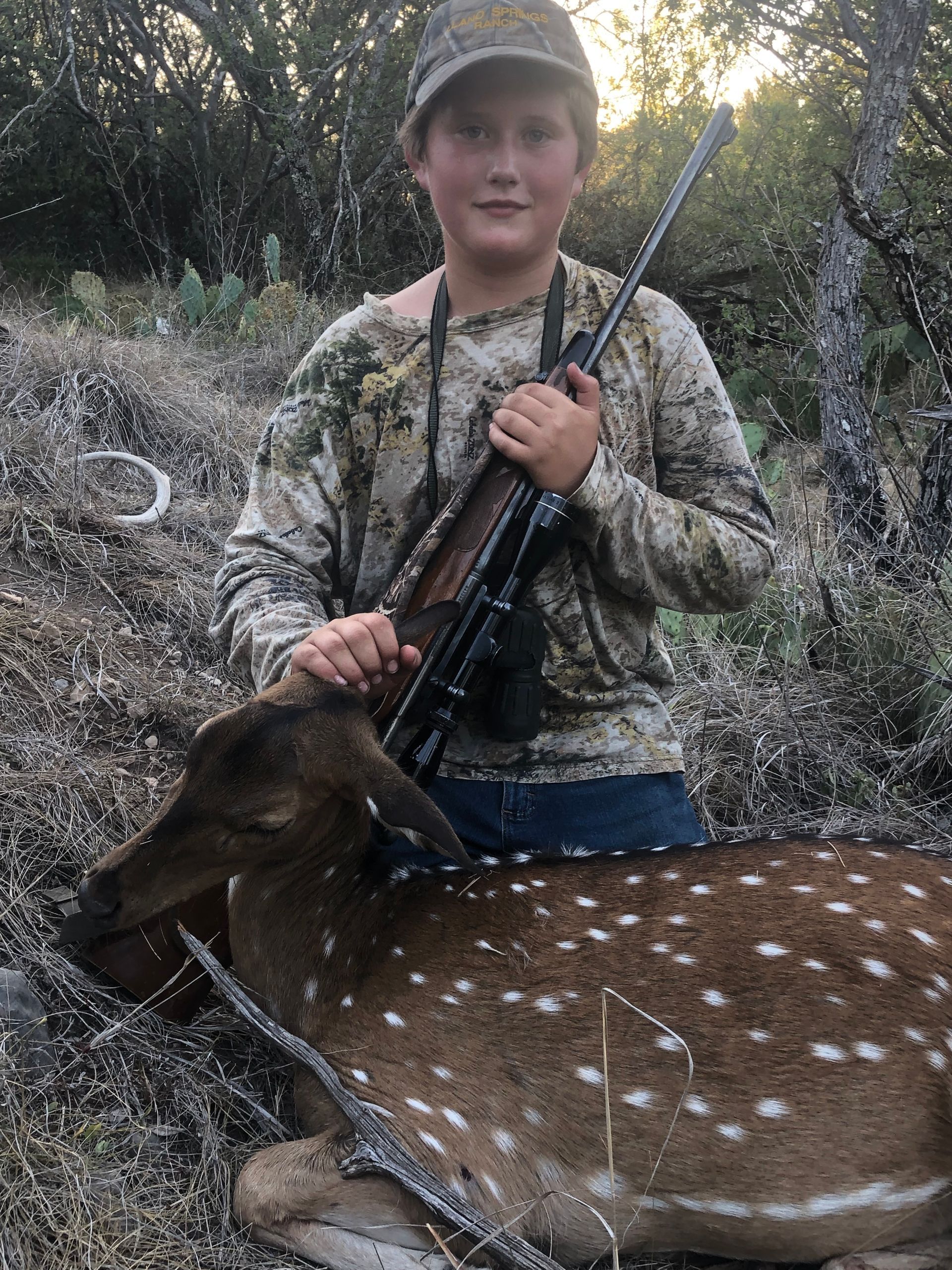 Boy in camouflage holds rifle, stands over a spotted deer in a wooded area.