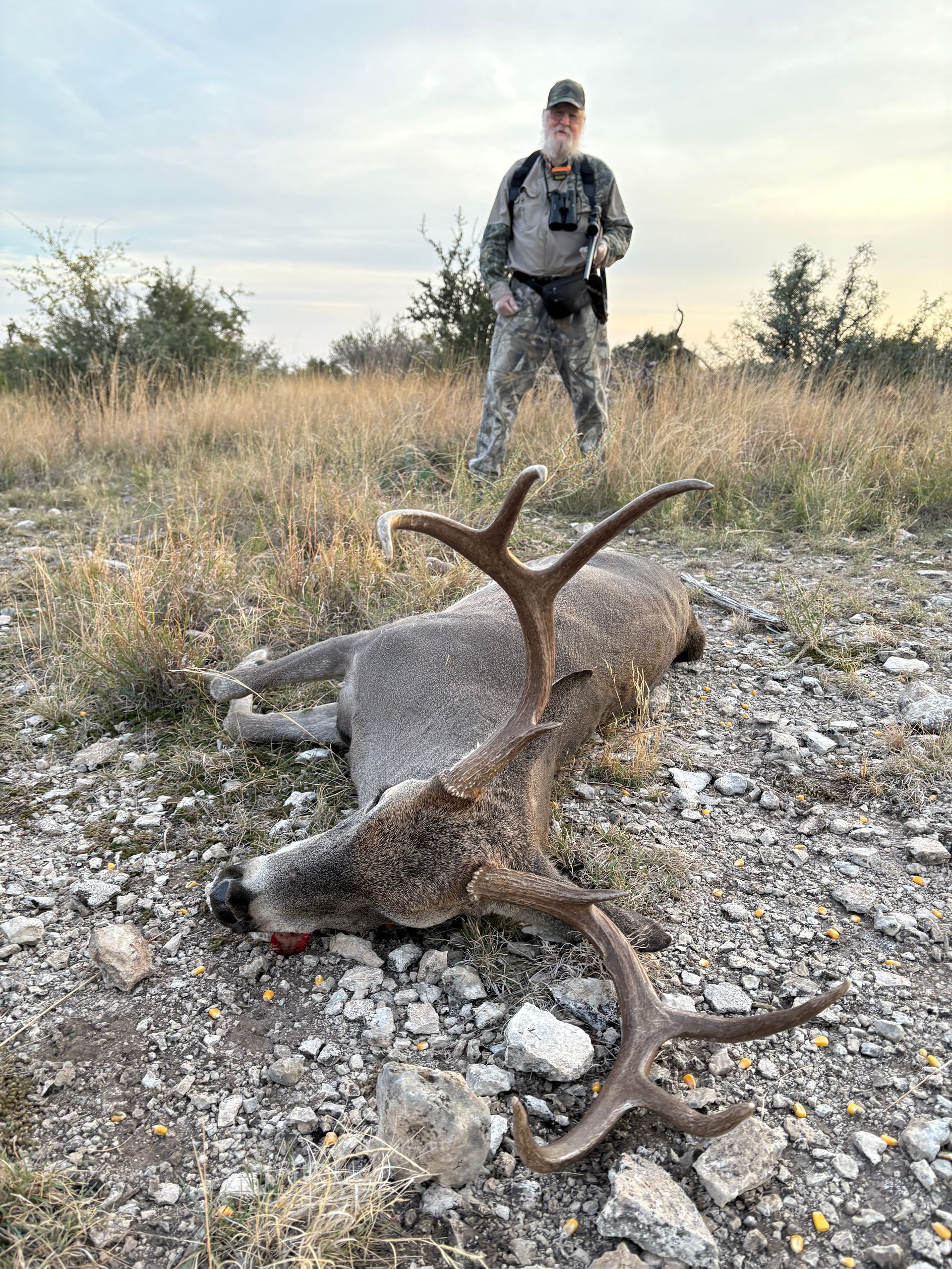 Hunter stands behind a harvested deer with large antlers in a field. Cloudy sky.