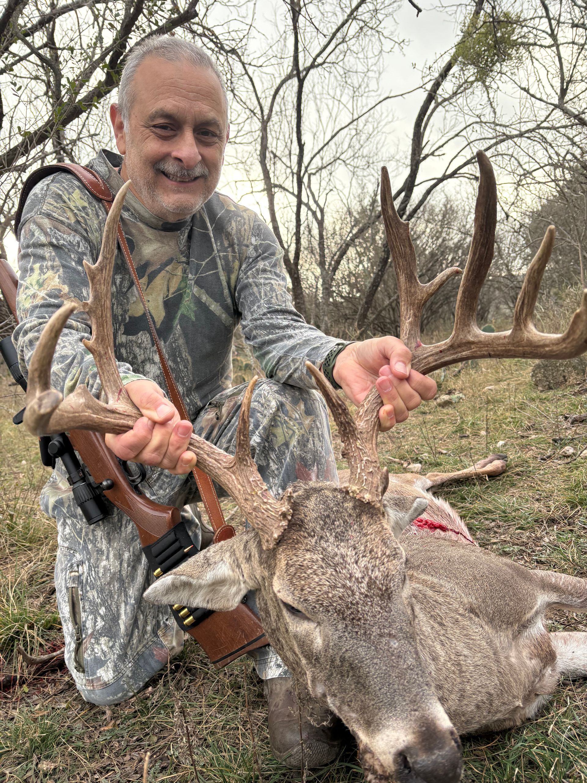 Man in camo kneels beside a harvested buck, holding antlers; outdoors.
