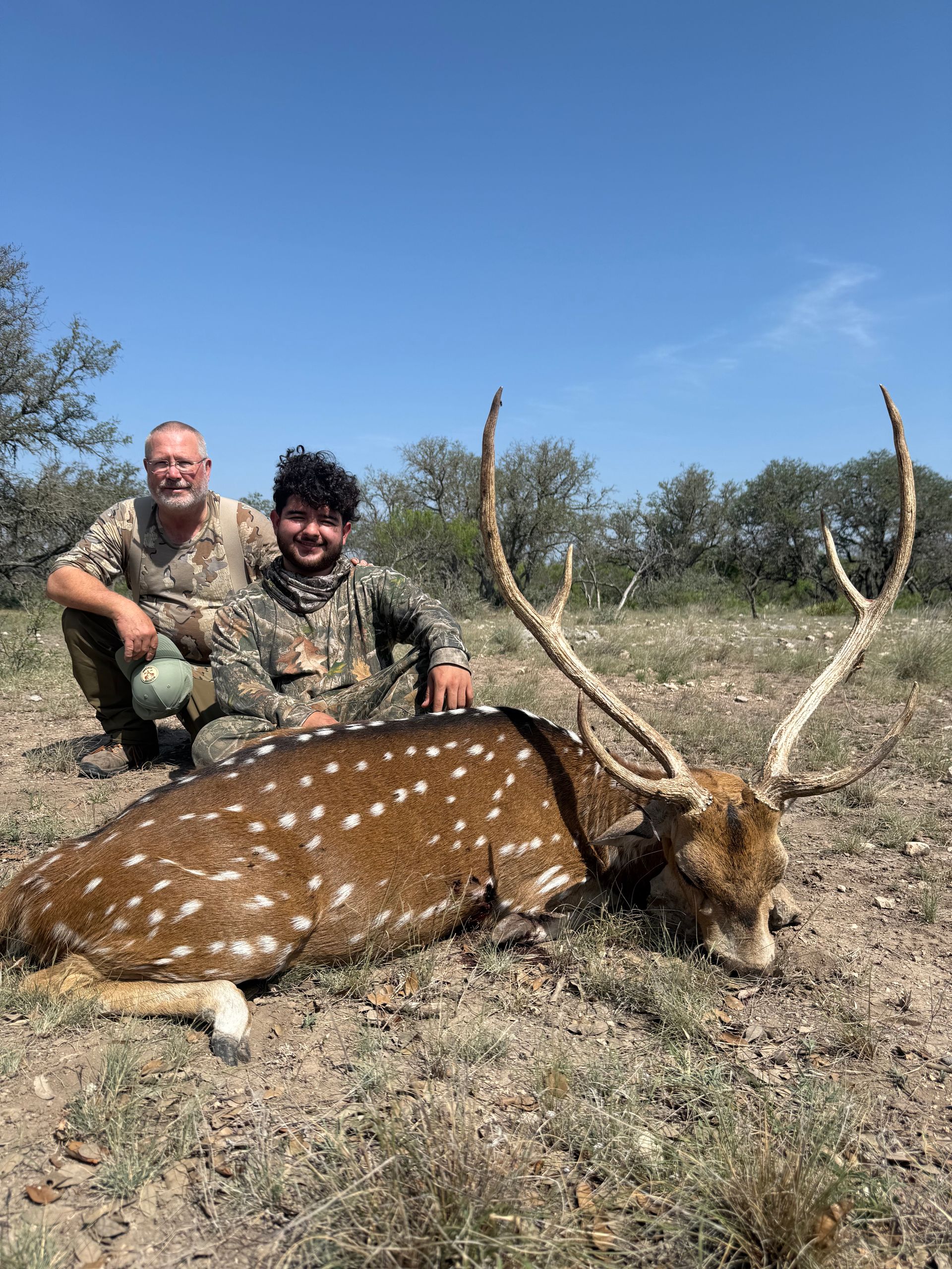Two people kneel beside a spotted axis deer lying on the ground in a field.