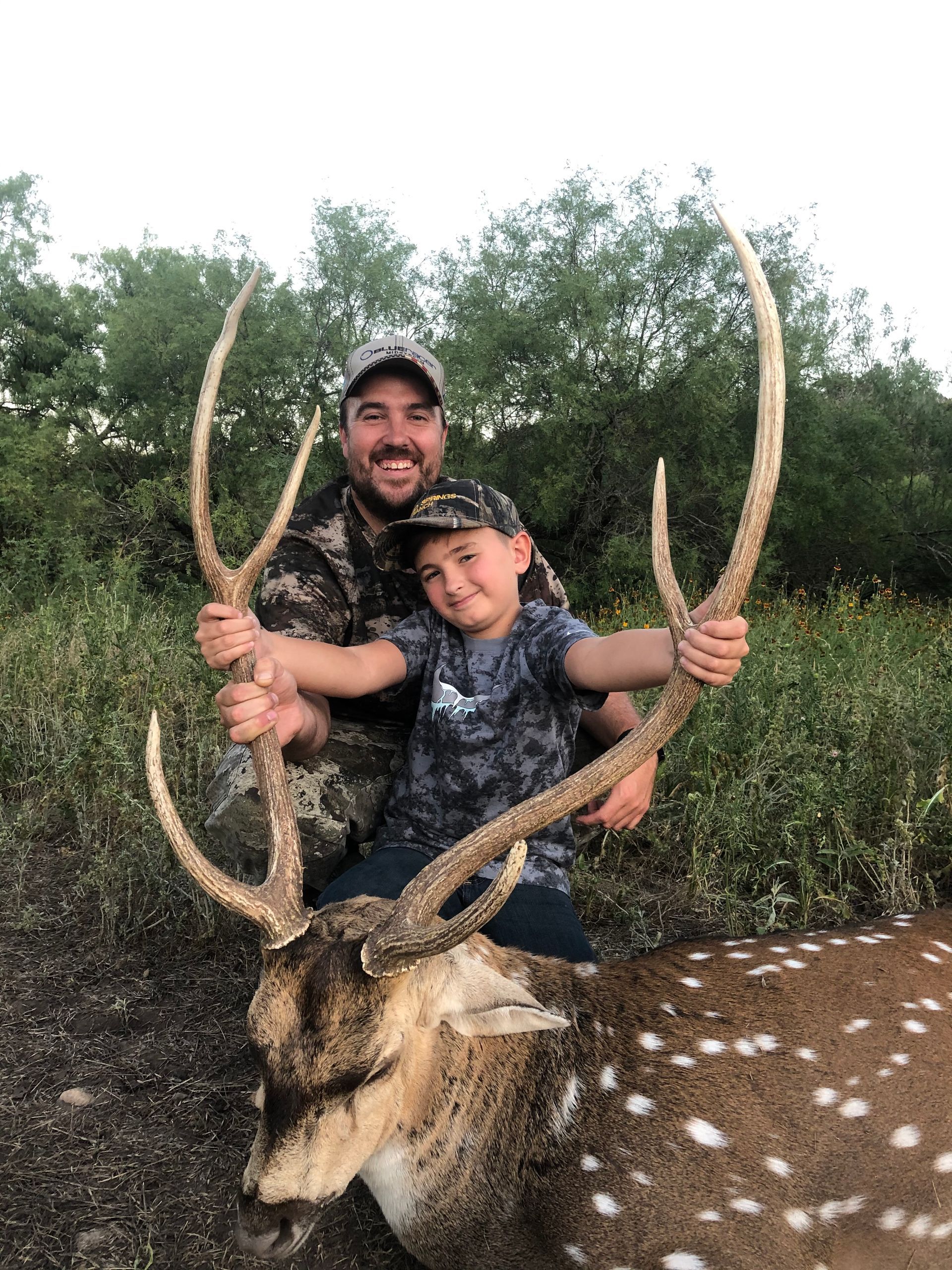 Man and child smiling, holding large antlers over a spotted deer they shot outdoors.