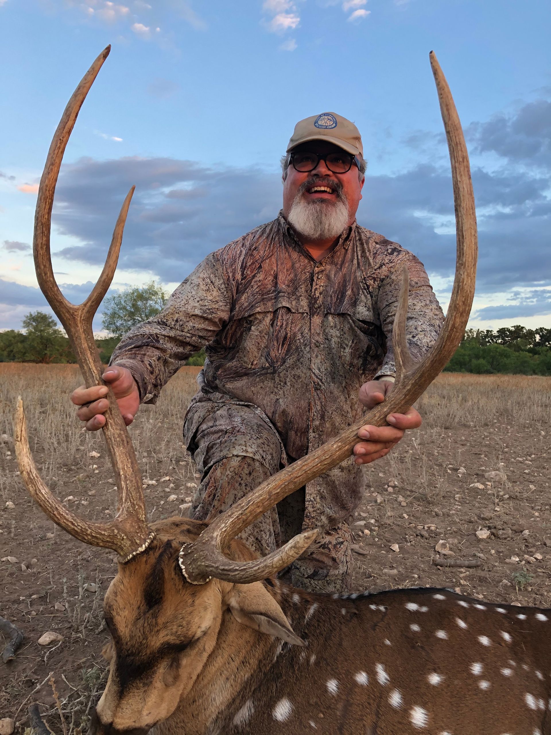Man kneels, holding large antlers of a spotted deer in a field. Smiling, wearing camo, and cap.