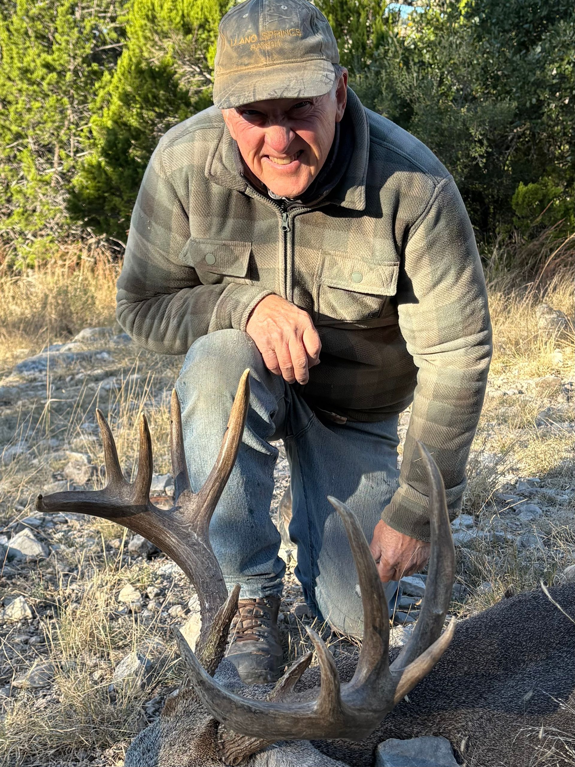 Man kneeling, holding large deer antlers, in outdoor setting. He's smiling.