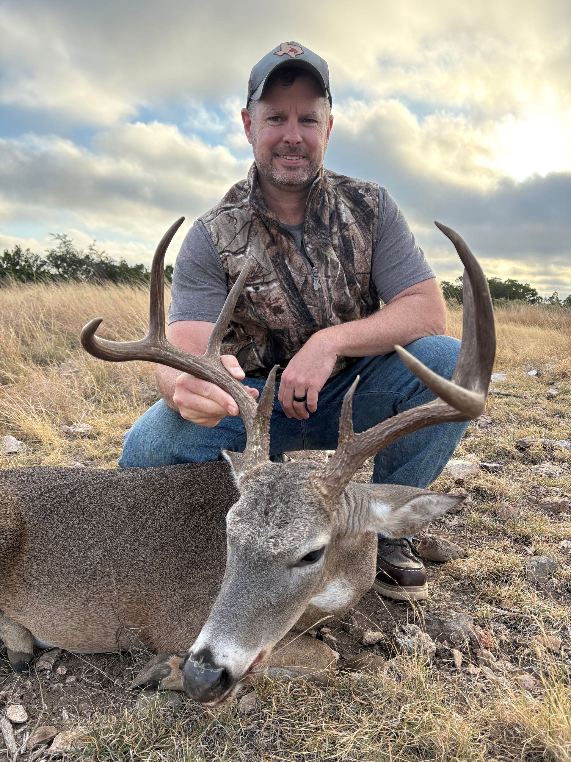 Man kneels beside a harvested deer in a field. He holds its antlers, smiling. Overcast sky.