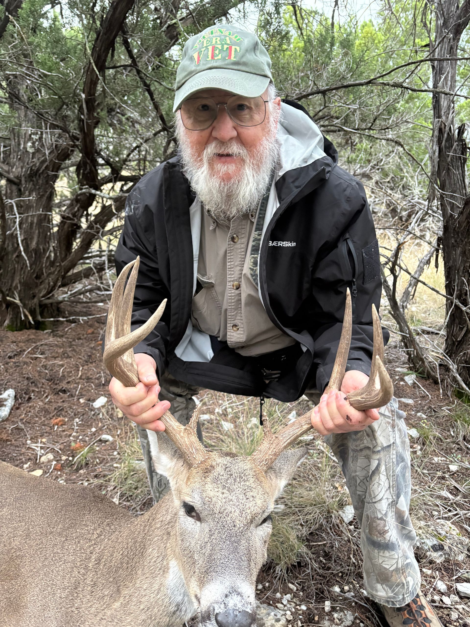 Man holding deer antlers outdoors, wearing a hat, beard, and jacket.